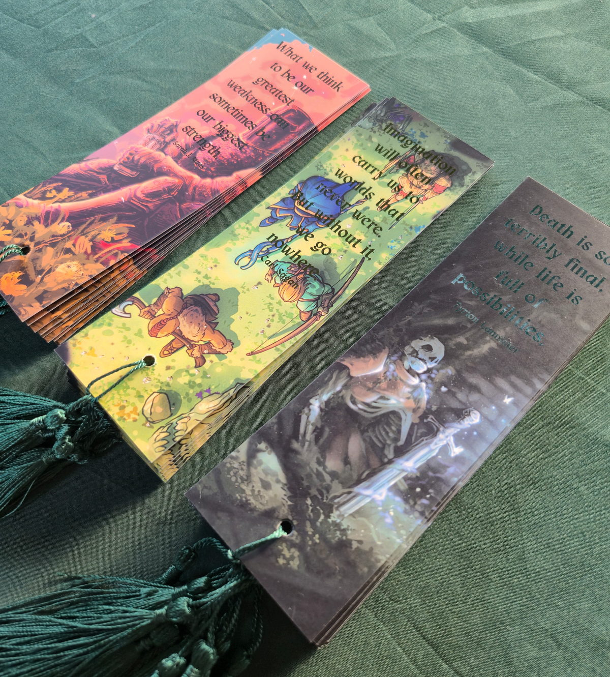 Three printed bookmarks with tassel ends laid on a green fabric surface. Each bookmark features fantasy artwork and a quote; one shows a character in armor, one shows mushrooms and a sword, and one shows a skeleton with a sword.